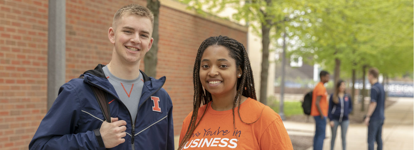 Two students stand underneath the trees outside of the Business Instructional Facility